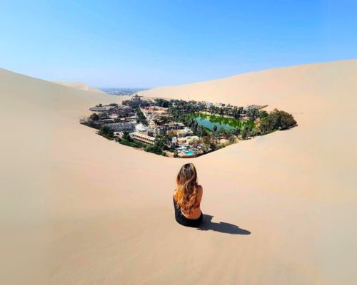 Woman sitting on a dune gazing at the Huacachina Oasis below.